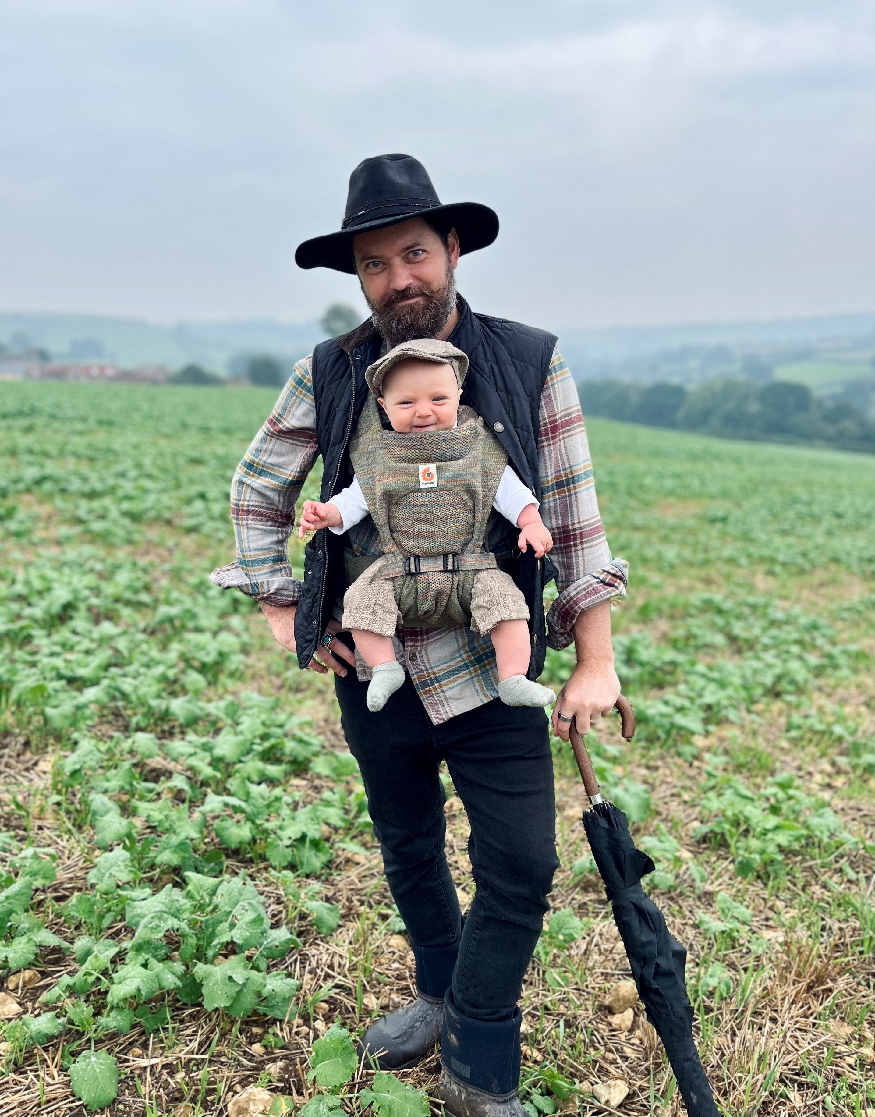 Jack Harris holding his baby in a carrier, standing in a green field—Tally Ho founder.
